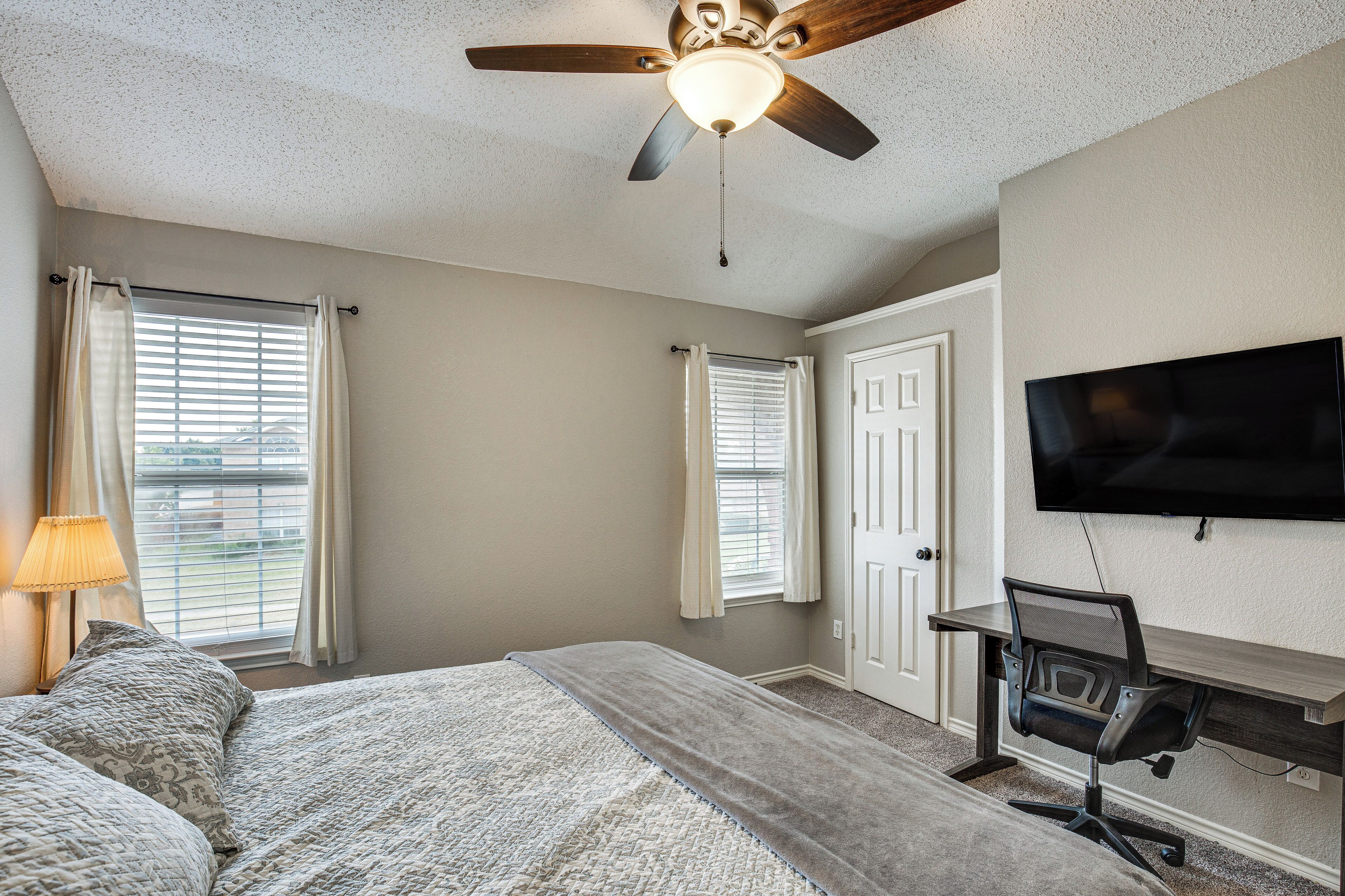 A bedroom with a bed, a desk, a chair, a ceiling fan, and a TV mounted on the wall.