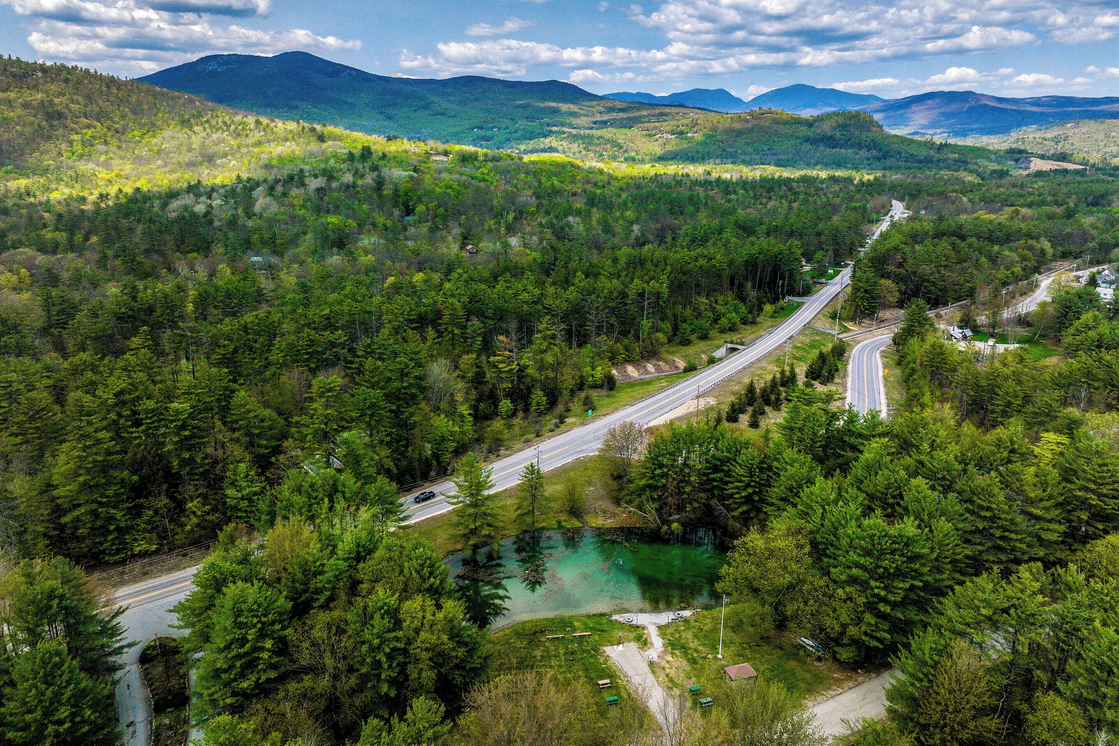 A winding road through a forested valley with a small lake and distant mountains.