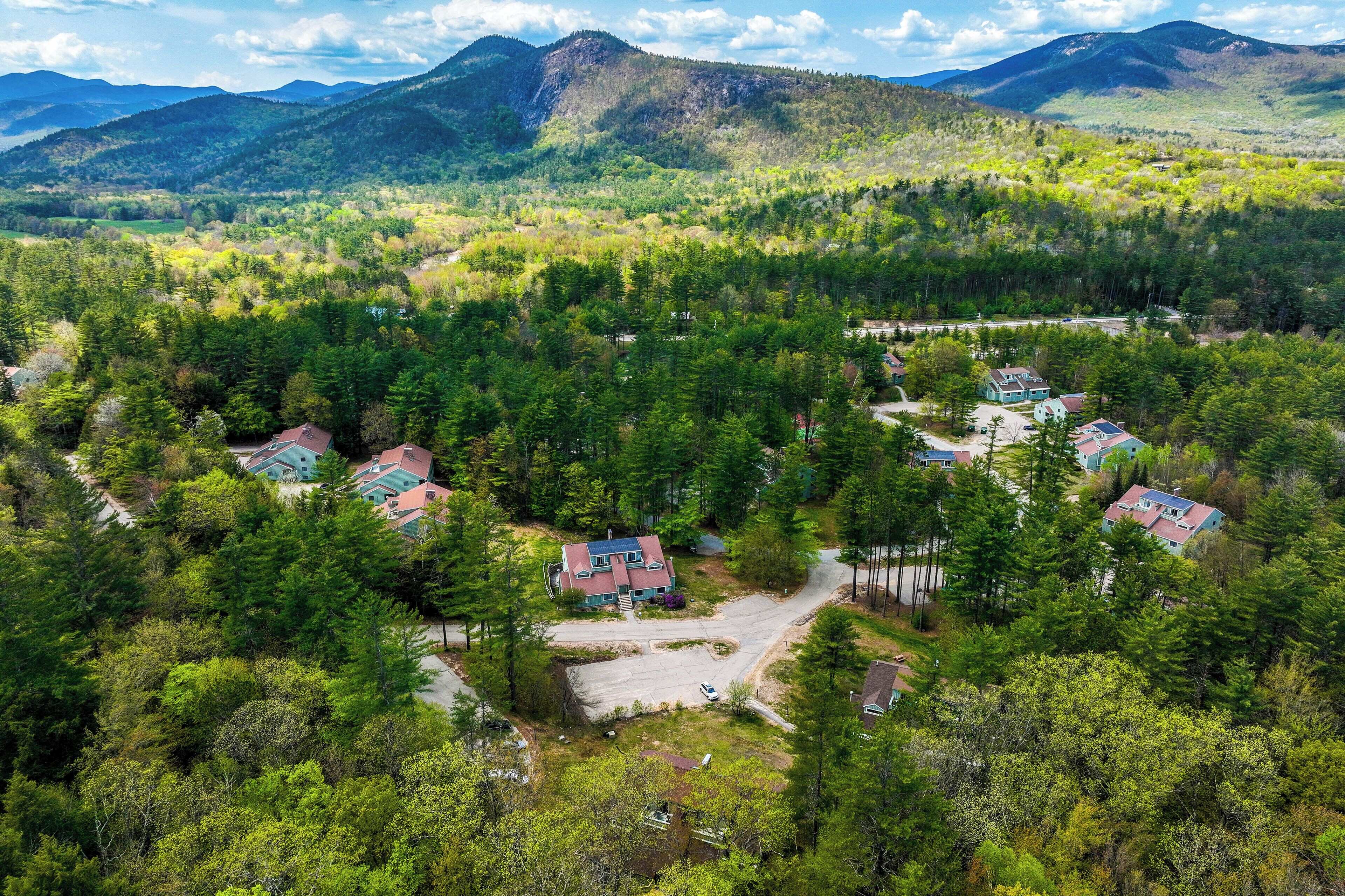 An aerial view of a residential area with houses surrounded by dense forest, a winding road, and mountains in the background.