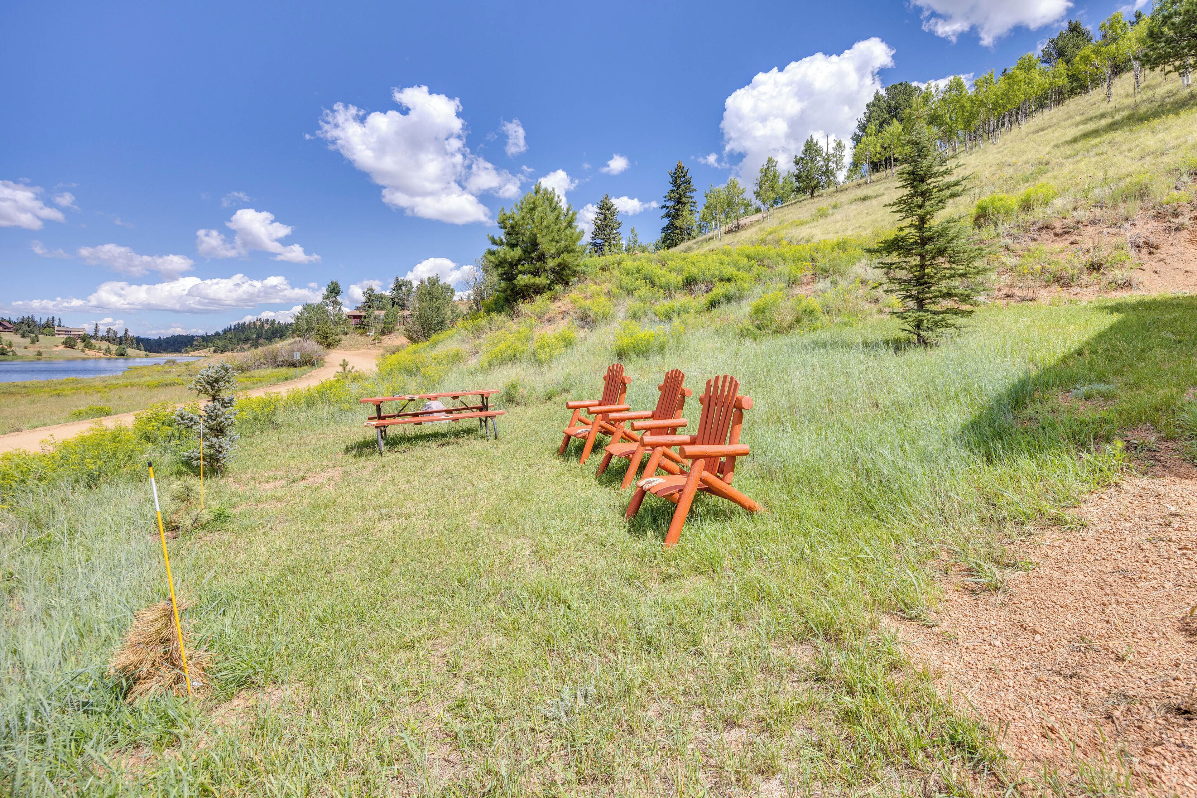 A picnic area with chairs and a table in a grassy field with a lake and trees in the background.