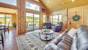 A spacious living room with a leather sofa, a coffee table, and a fireplace. There are large windows and a view of the outdoors. - Lakefront Divide Cabin w/ Game Room! (Woodland Park)