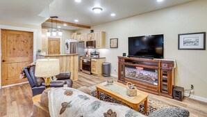 A living room with a fireplace, a television, and a kitchen area with wooden cabinets and appliances. - Cozy Eureka Getaway - Hike, Bike, Hunt & Fish! (Eureka)