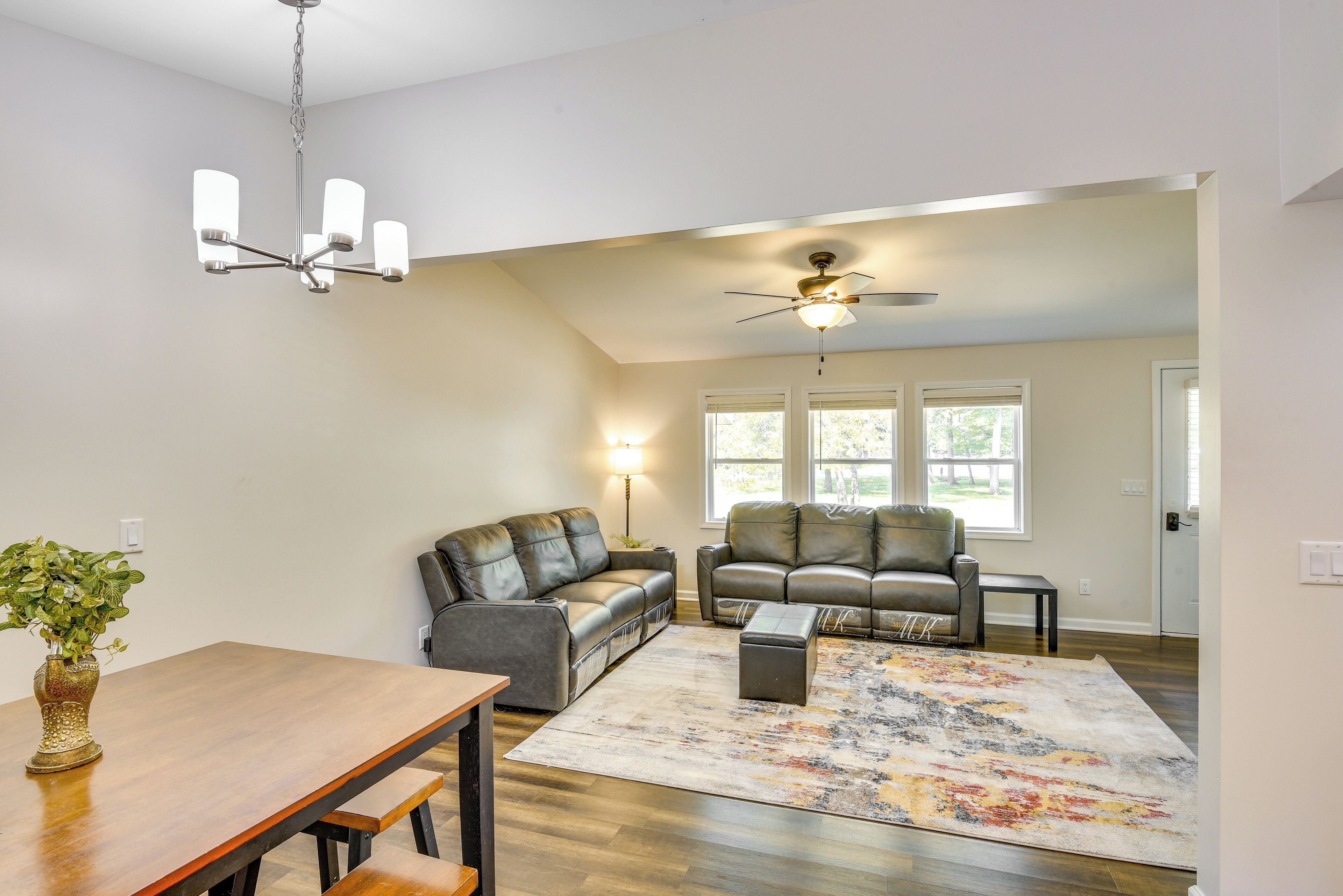 A spacious living room with a wooden dining table, two leather sofas, a ceiling fan, and a large window providing natural light.