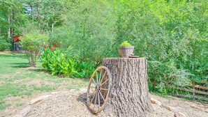 A rustic outdoor setting with a wooden wheel planter on a tree stump, surrounded by lush greenery and a wooden fence. - Cabin w/ Fire Pit - 7 Mi to Downtown Carrollton! (Carrollton)