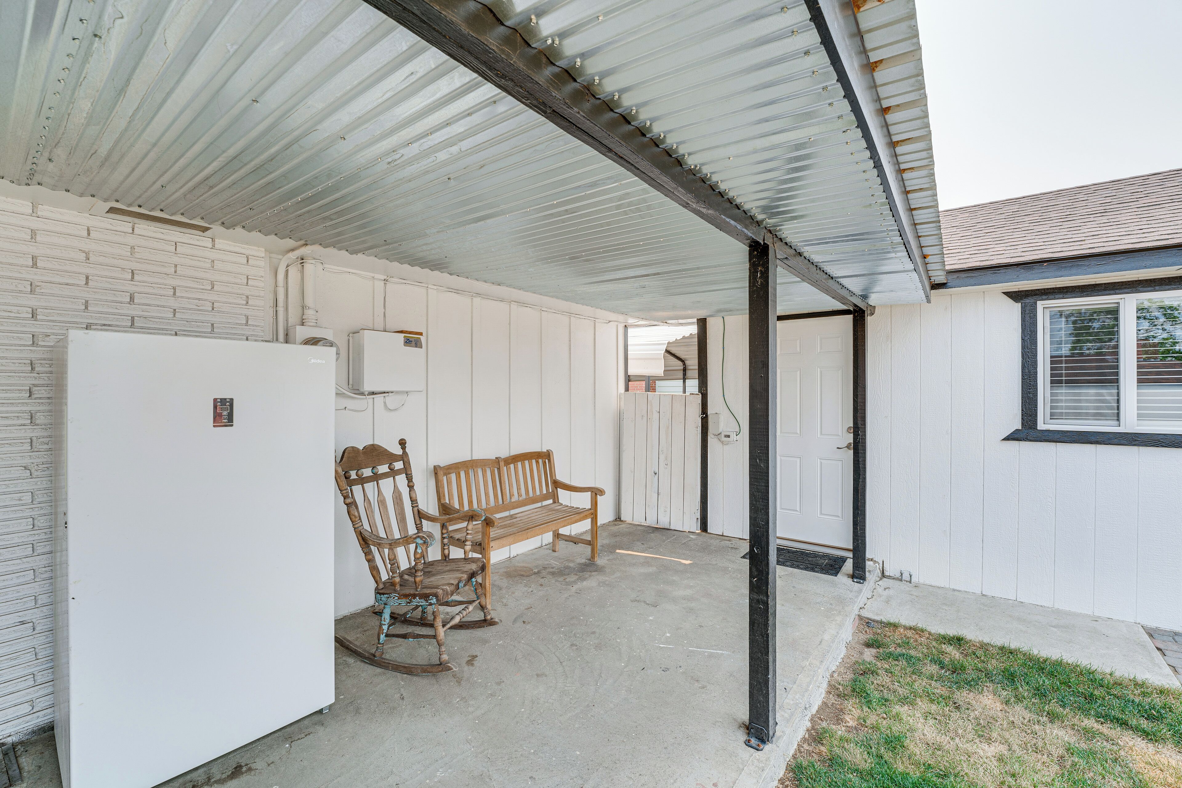 A covered outdoor area with a white refrigerator, a wooden bench, and a rocking chair.