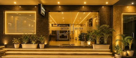 L’entrée d’un hôtel, avec une enseigne indiquant « DECENT SUITES », un comptoir d’accueil et des plantes en pot.