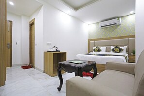 A hotel room with a bed, a bedside table, a chair, a small table, and an air conditioning unit. - Hotel Decent Suites - Delhi Airport (New Delhi)