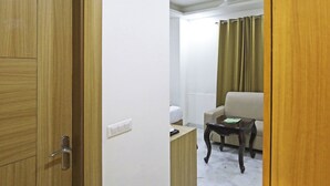 Habitación de hotel con puerta de madera, pared blanca, ventilador de techo, sofá color beige, pequeña mesa de madera y cortina verde.