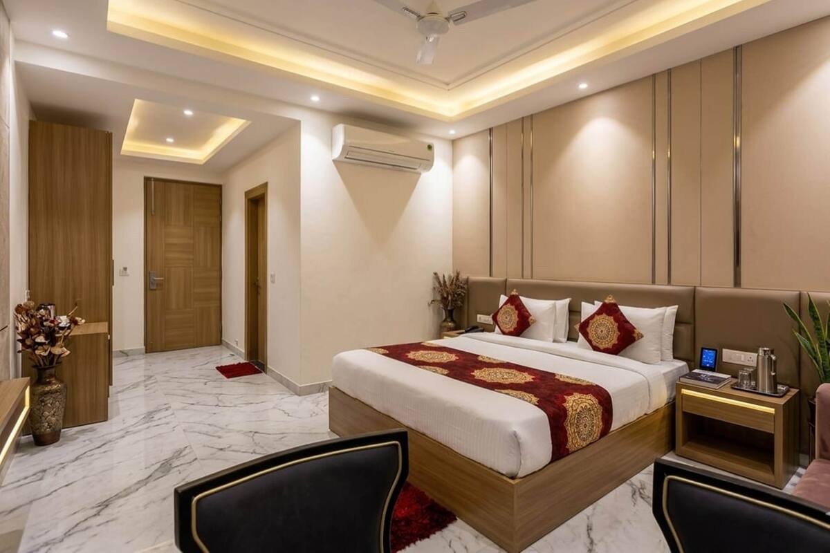 A modern hotel room with a large bed, a desk, a chair, a bedside table, and an air conditioning unit.