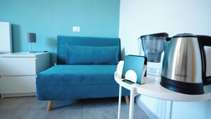 Une chambre moderne avec un canapé de couleur turquoise, une table d’appoint blanche sur laquelle se trouvent une bouilloire et un récipient en verre, et une table de chevet blanche avec une lampe.