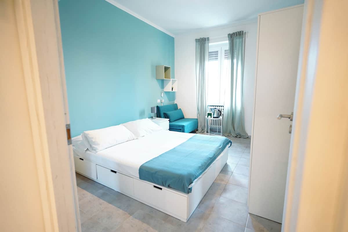 A bedroom with a bed, a nightstand, a chair, a small table, and a window with curtains.