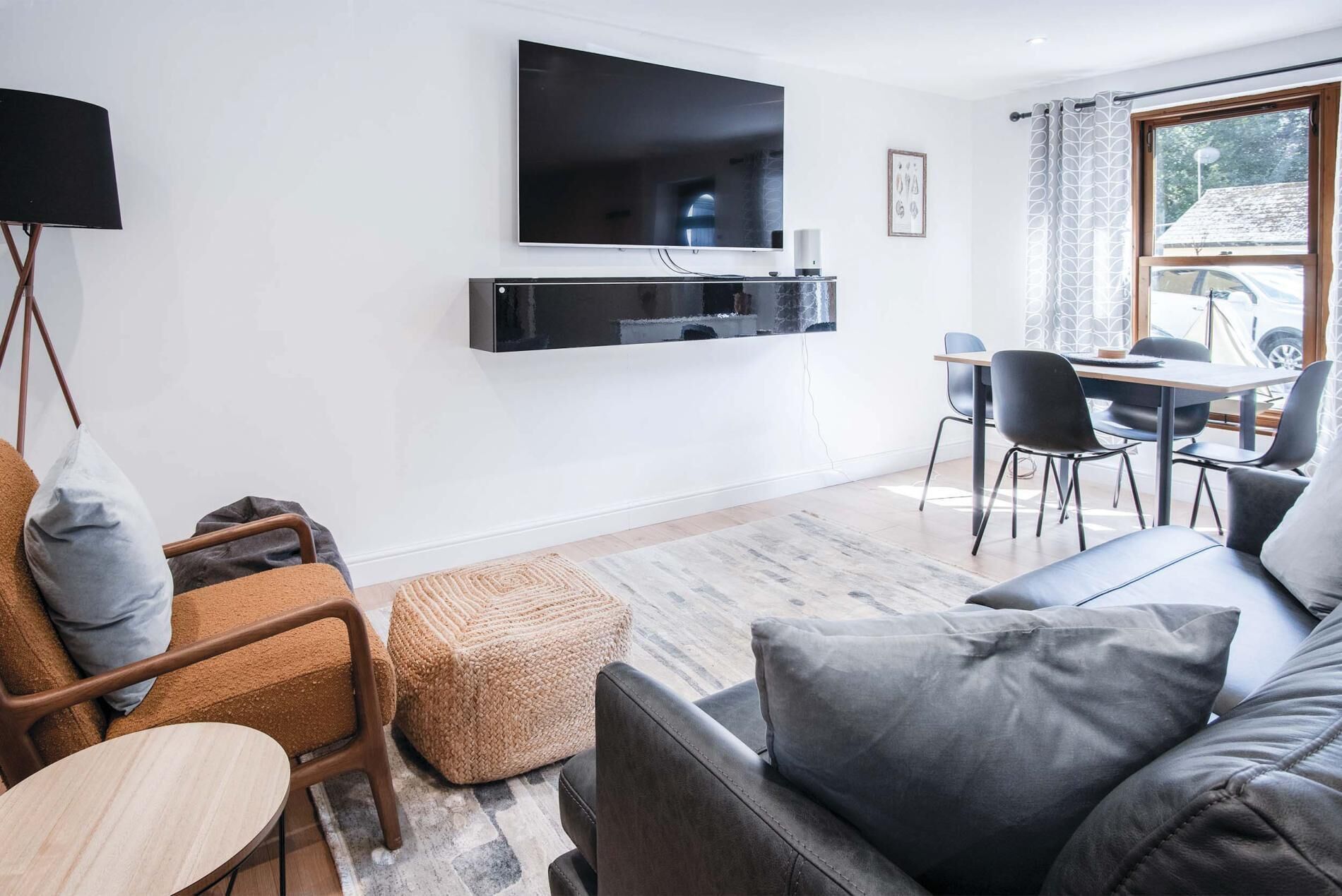 A modern living room with a flat-screen TV mounted on the wall, a dining area with a table and chairs, and a large window with curtains.