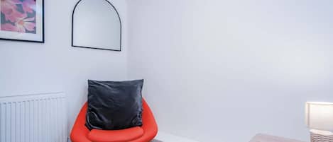 Una habitación moderna con una silla roja, un cojín negro, un radiador blanco y una pequeña mesa de madera con una lámpara.