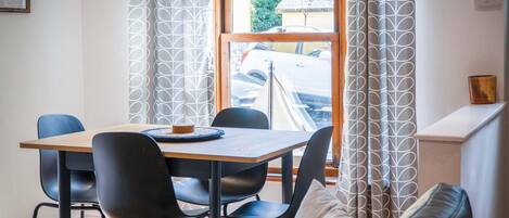 Un coin repas avec une table en bois et quatre chaises, une fenêtre avec des rideaux et une vue sur une voiture garée à l’extérieur.
