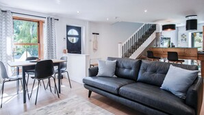 Una sala moderna con un sofá de cuero oscuro, un comedor con mesa y sillas, una escalera y una cocina con barra.