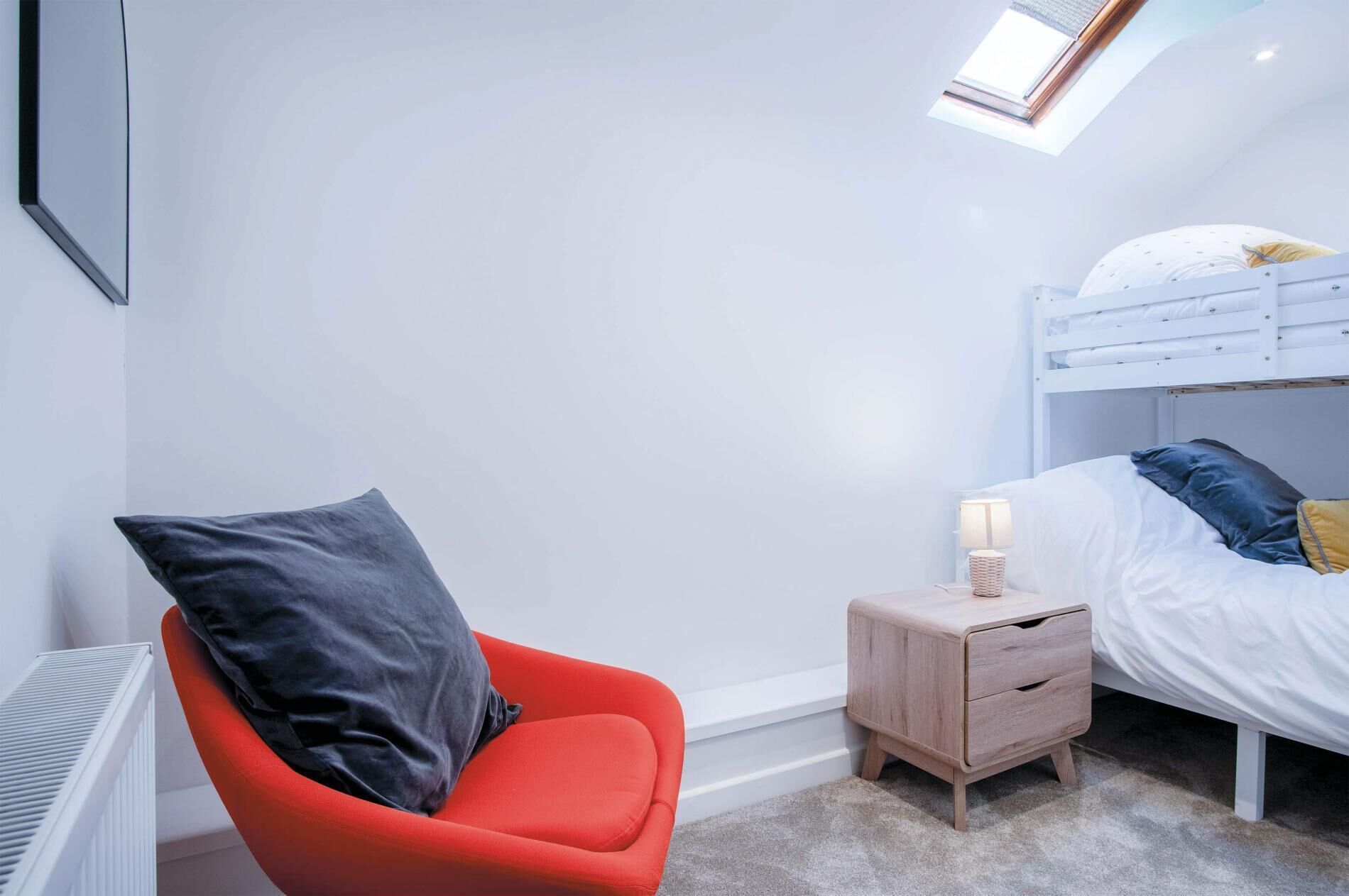 A bedroom with a bunk bed, a red chair, a bedside table with a lamp, and a radiator.