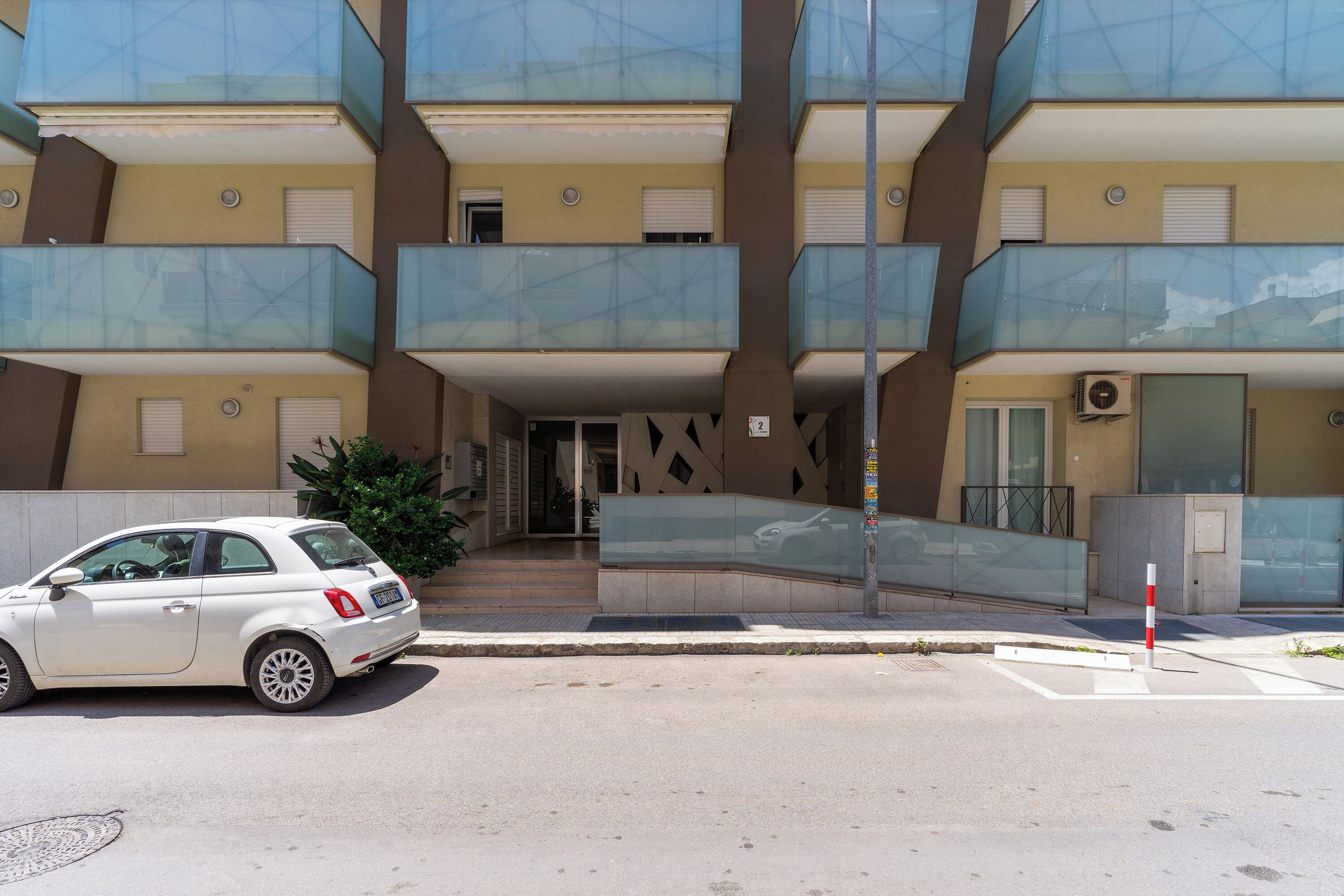Un edificio residencial de varios pisos con balcones, un automóvil estacionado al frente y una entrada despejada.