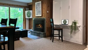 A cozy room with a fireplace, wooden walls, a dining area, and a view of trees through the window.