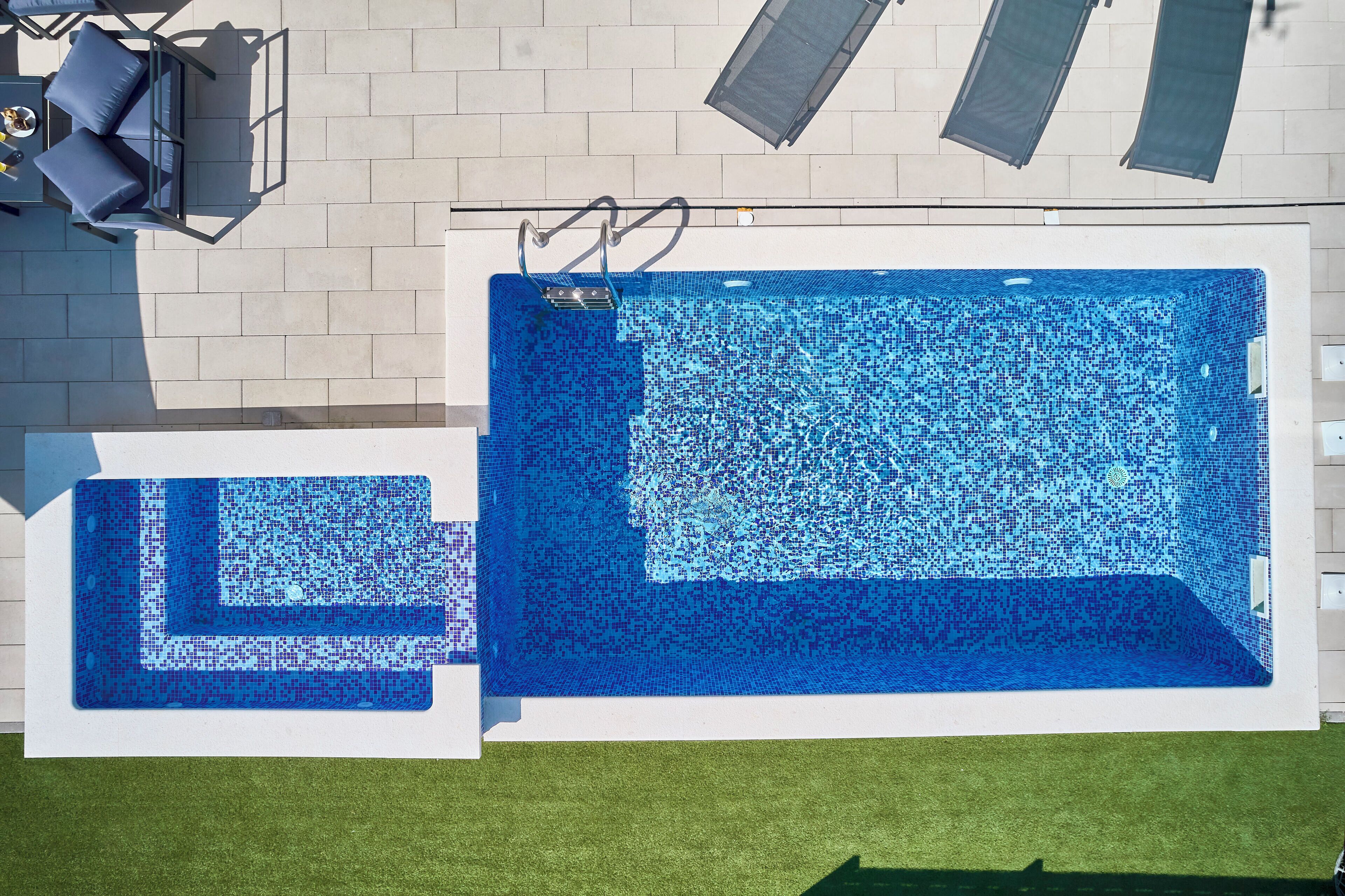 A rectangular swimming pool with a small adjacent pool, surrounded by a tiled deck and a grassy area.