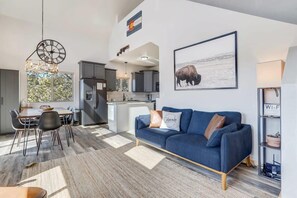 Een moderne woonkamer met een blauwe bank, een eethoek met een tafel en stoelen, een keuken met grijze kastjes, en een groot raam.