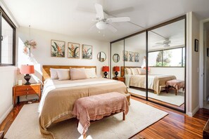A bedroom with a large bed, a bedside table with a lamp, a mirror, and a window with a view of greenery. - Kona Makai#1-102: Nearly Oceanfront, Ground Floor, with AC & Newly Remodeled! (Kailua Kona)