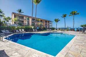 A swimming pool with lounge chairs and palm trees, surrounded by a building with balconies. - Kona Makai#1-102: Nearly Oceanfront, Ground Floor, with AC & Newly Remodeled! (Kailua Kona)