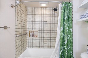 A shower area with a white bathtub, a green shower curtain, and a shelf with toiletries. - Kona Makai#1-102: Nearly Oceanfront, Ground Floor, with AC & Newly Remodeled! (Kailua Kona)