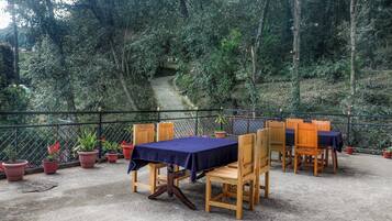 An outdoor patio with wooden tables and chairs, surrounded by potted plants and a view of a forested area.