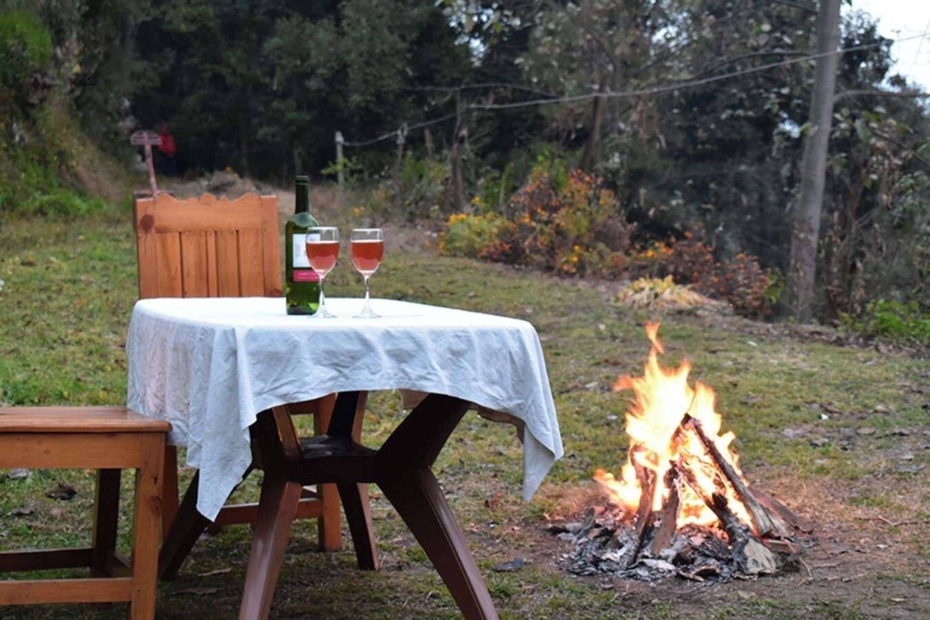 Una mesa al aire libre con mantel blanco, dos copas de vino tinto y una botella de vino. Un fuego arde al fondo.