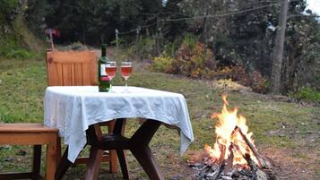 Una mesa al aire libre con mantel blanco, dos copas de vino tinto y una botella de vino. Un fuego arde al fondo.