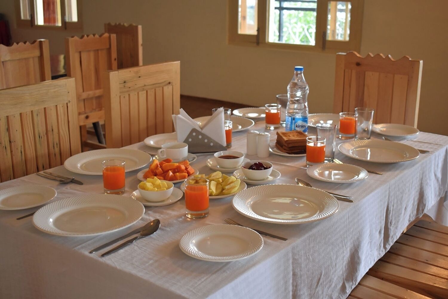 Une table de petit-déjeuner dressée avec des assiettes, des couverts et une variété d’aliments pour le petit-déjeuner, notamment des fruits, du jus et une bouteille d’eau.