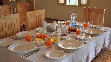 Une table de petit-déjeuner dressée avec des assiettes, des couverts et une variété d’aliments pour le petit-déjeuner, notamment des fruits, du jus et une bouteille d’eau.