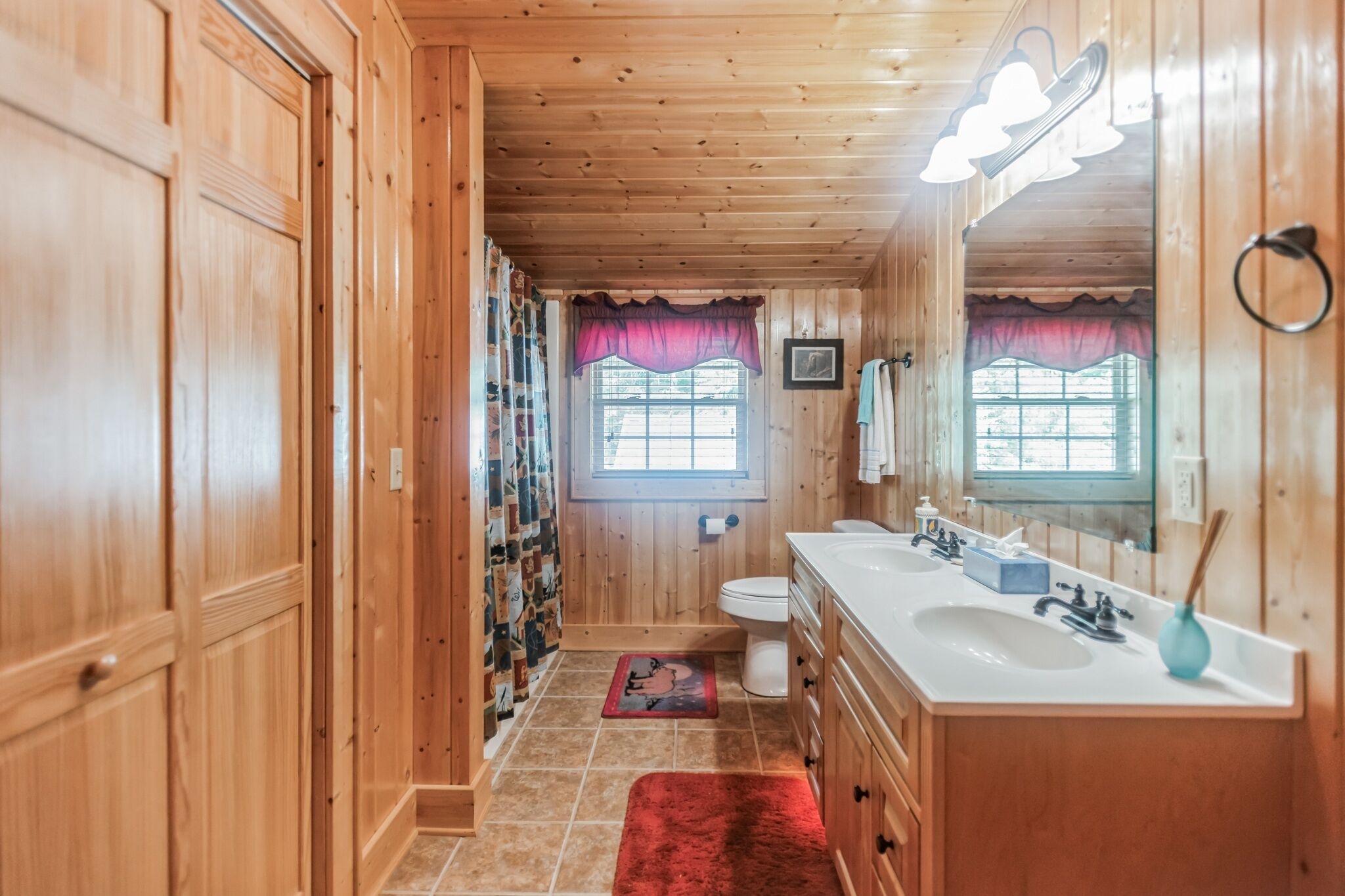 A wooden-paneled bathroom with a double sink, vanity, and a window with a curtain.
