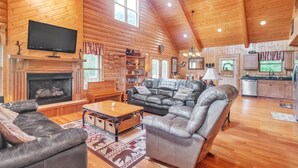 A spacious living room with a fireplace, leather sofas, a television mounted on the wall, and a kitchen area in the background.