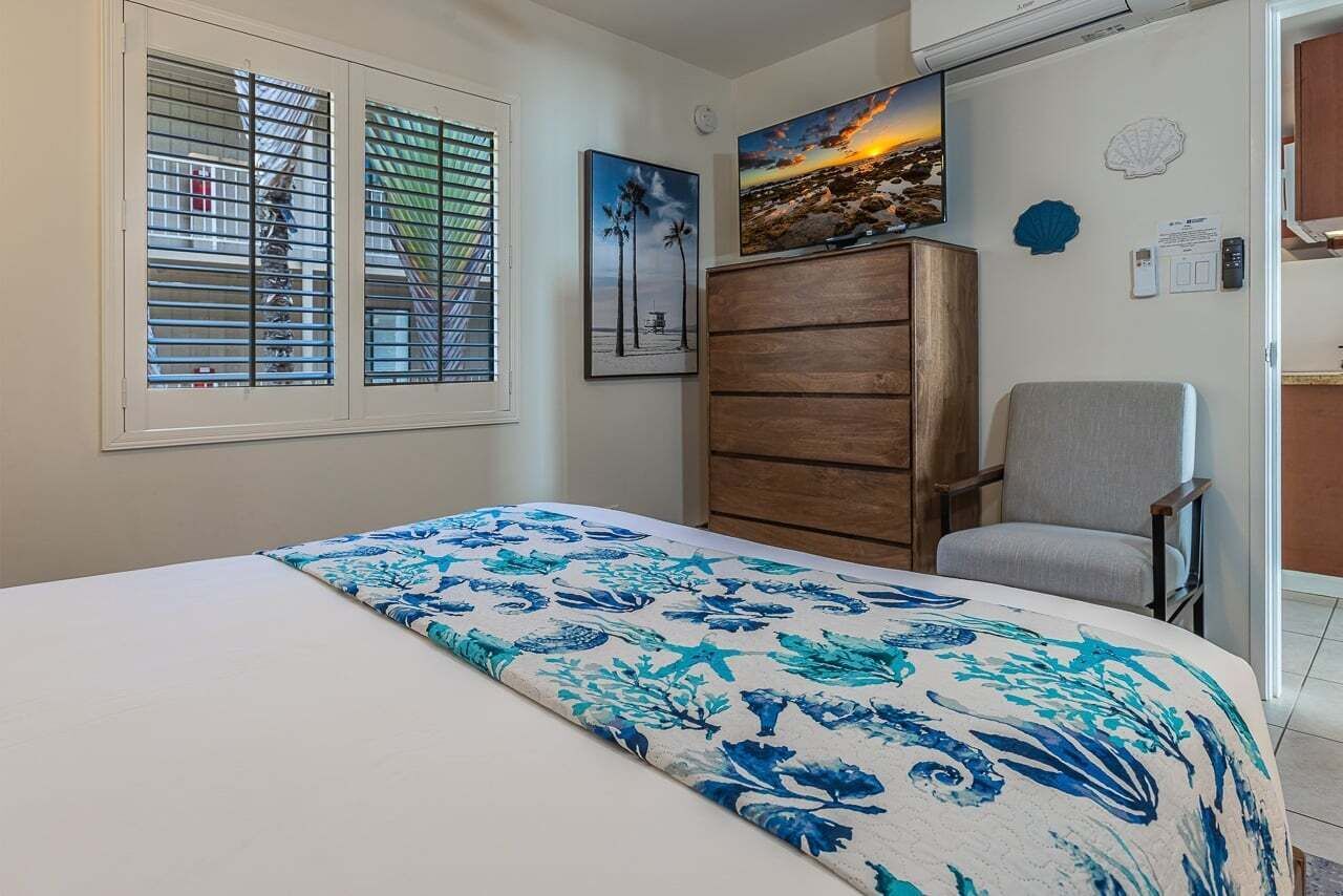 A bedroom with a bed, a chair, a wooden dresser, a window with blinds, and wall decorations including a sunset painting and a shell.