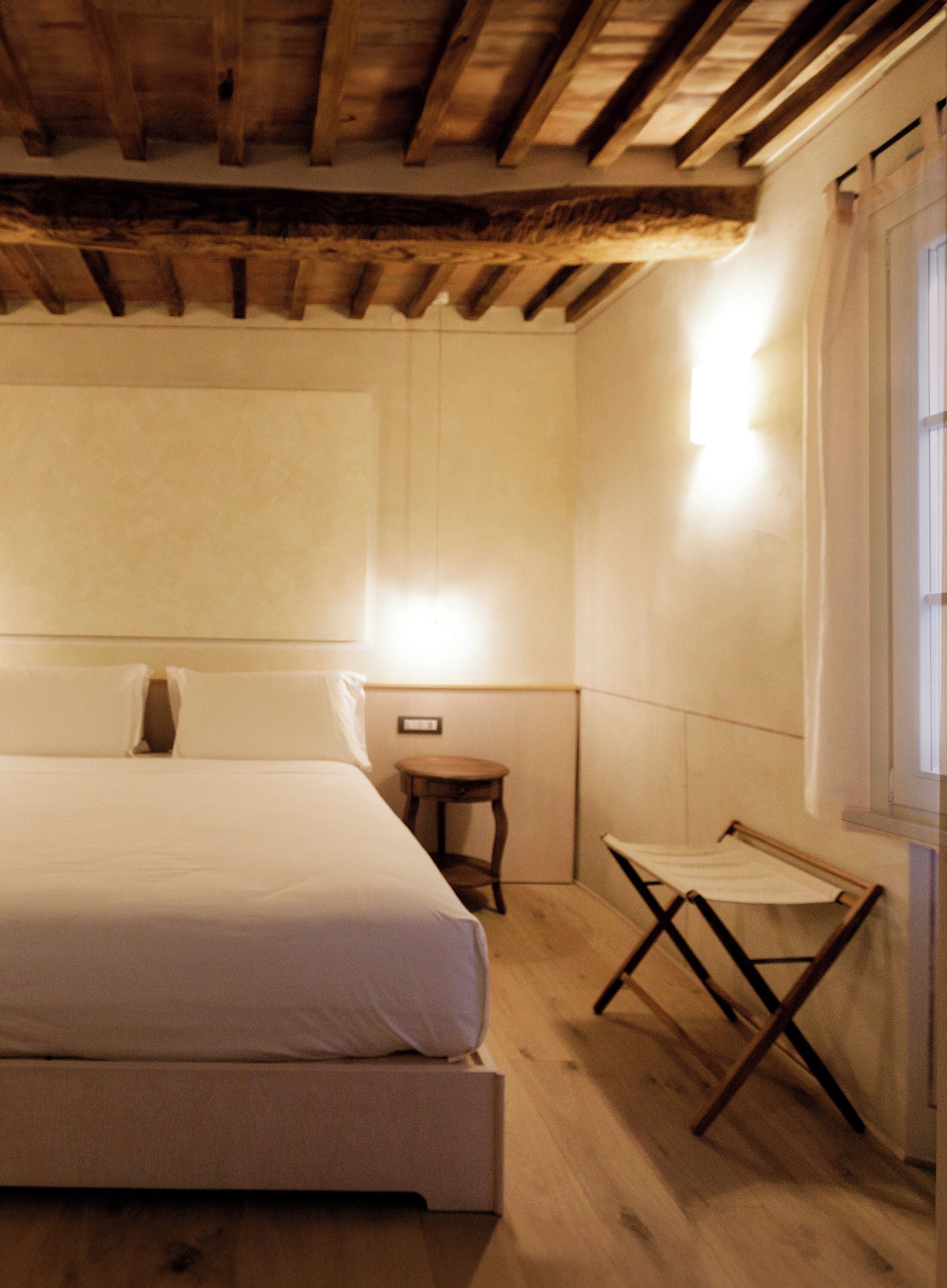 Une chambre à coucher avec un lit, un petit tabouret en bois et une fenêtre avec des rideaux.