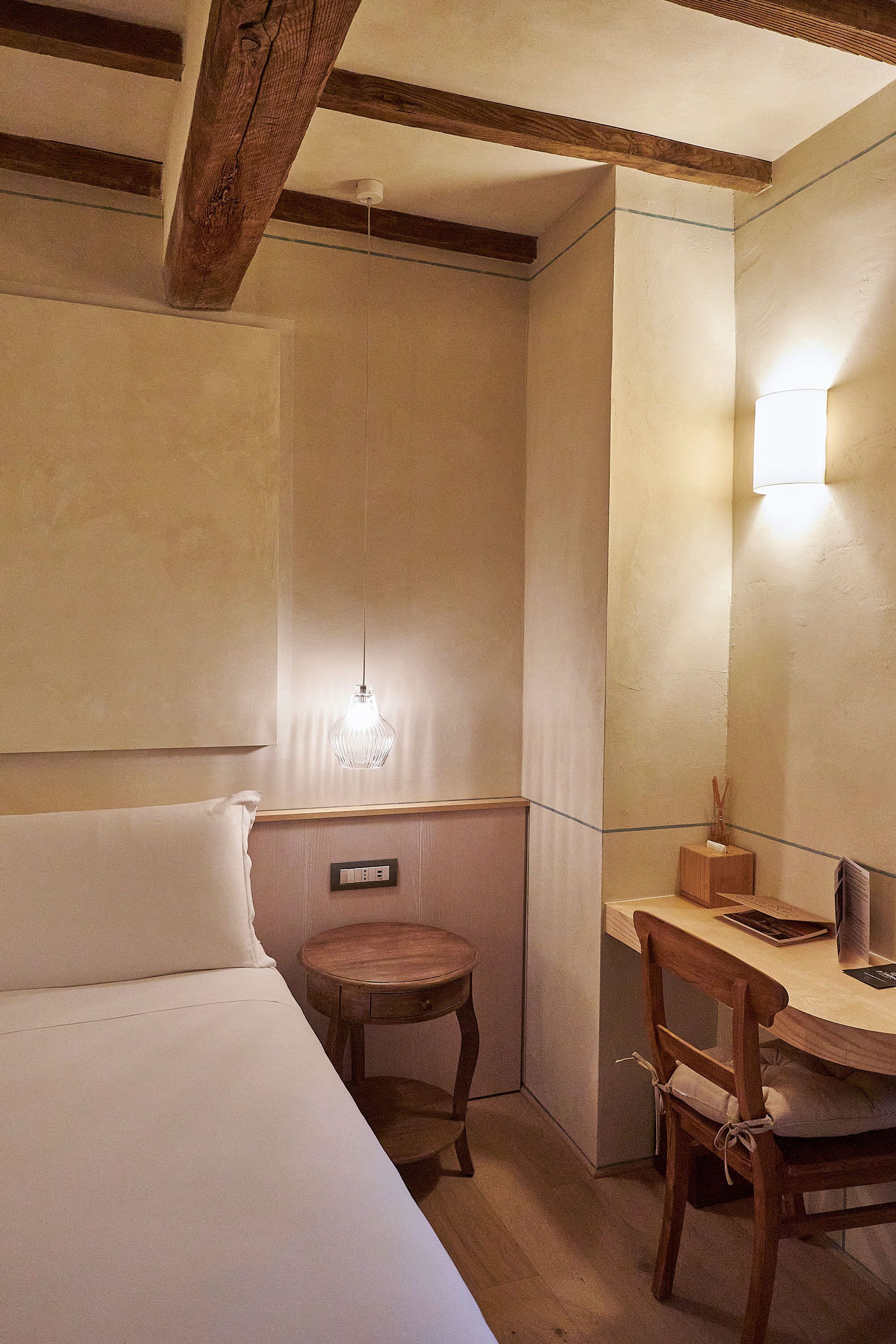 Ein Zimmer mit einem Bett, einem kleinen Holztisch, einem Stuhl, einer Wandleuchte und einem Deckenventilator.