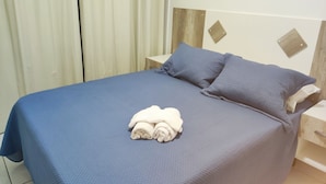 Un letto ben rifatto con copriletto blu e due cuscini, un asciugamano bianco piegato sul letto e una testiera in legno.