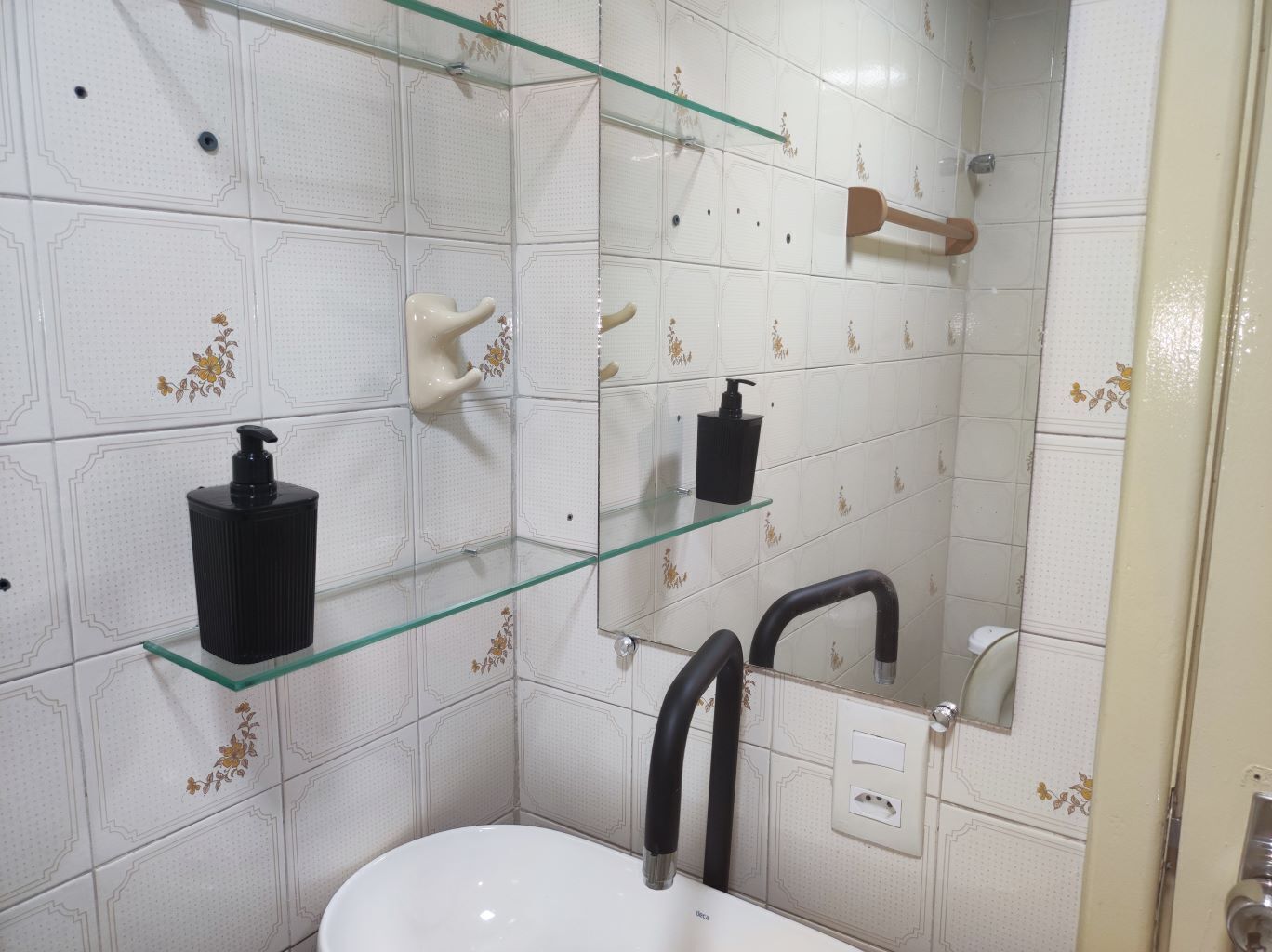 Un baño con paredes de azulejos blancos, un estante de vidrio con artículos de aseo, un lavamanos blanco y un espejo.