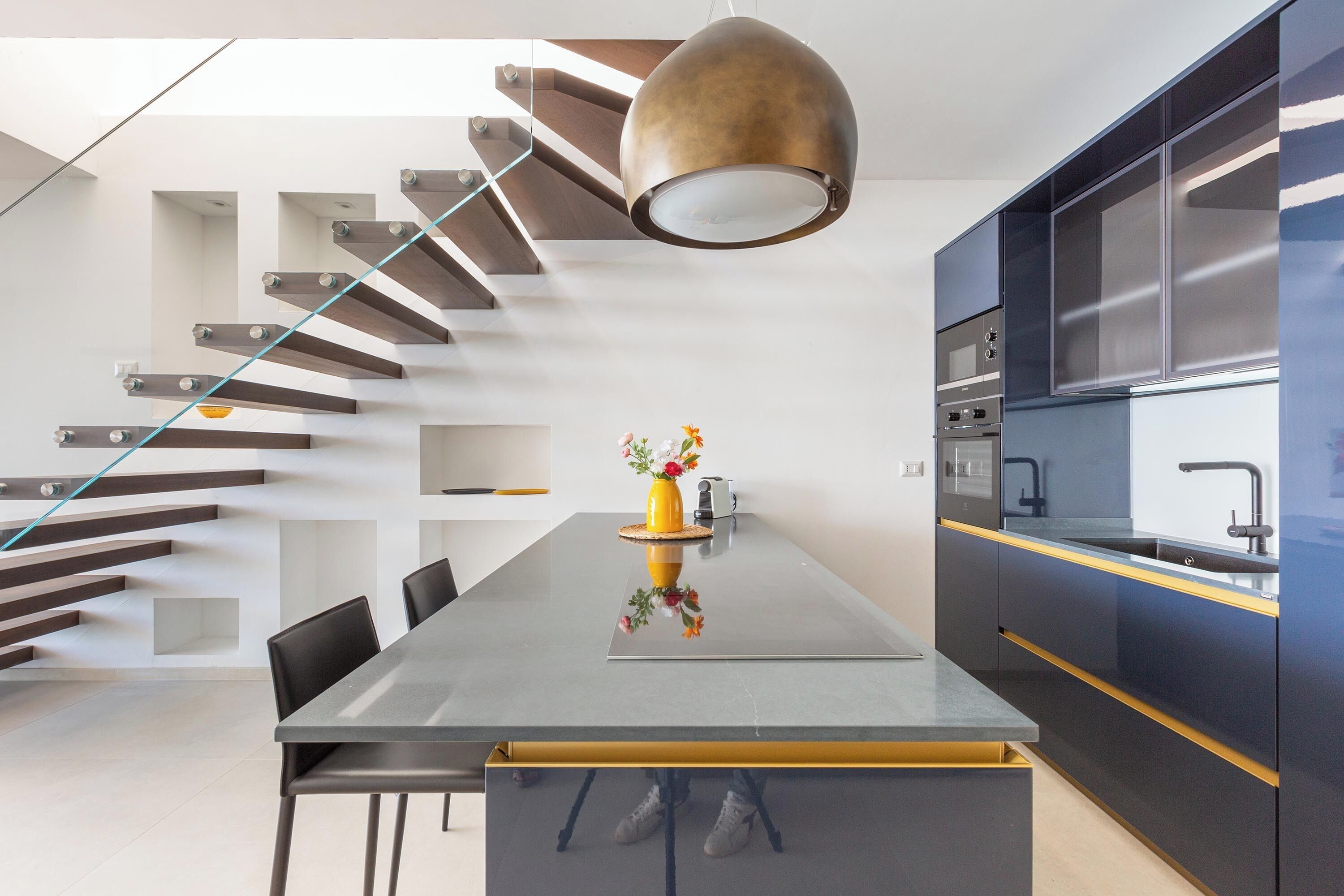 A modern kitchen with a central island, a staircase with glass railings, and built-in appliances.