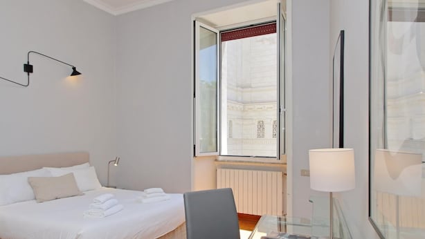A bedroom with a bed, a desk with a lamp, a window, and a view of a building. - Exclusive Historic Ghetto Flatinrome (ROMA)