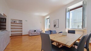 A modern living room with a wooden dining table, grey chairs, a light grey sofa, and a large window providing natural light. - Exclusive Historic Ghetto Flatinrome (ROMA)