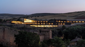 Un edificio moderno de varios niveles y grandes ventanales, rodeado de colinas ondulantes y algunos árboles.