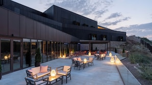 Un patio al aire libre con asientos y mesas, un edificio moderno con grandes ventanales y una pared de piedra al fondo.