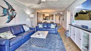 A modern living room with a blue sofa set, a coffee table, and a ceiling fan. - ☀️Land's End 3-403🐬Bay Views🏖️Beach Access on Grounds🌴Large Pool😎Top Floor (Treasure Island)