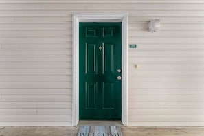 Una puerta verde con marco blanco, felpudo y placa con el número de la casa.
