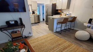A modern living room with a flat-screen TV, a wooden coffee table with decorative items, a kitchen area with a fridge, microwave, and bar stools, and a cozy seating area with a round ottoman.