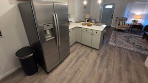 A modern kitchen with a stainless steel refrigerator, a water dispenser, and a built-in oven. There is a small trash bin and a wooden floor.
