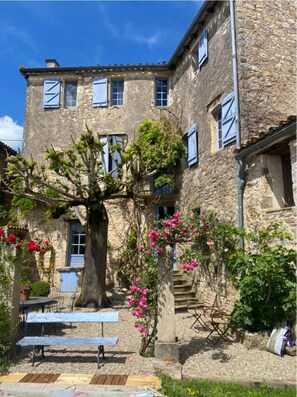 Relax in the serene courtyard surrounded by a large tree and blooming flowers, with benches and a table for a peaceful retreat.