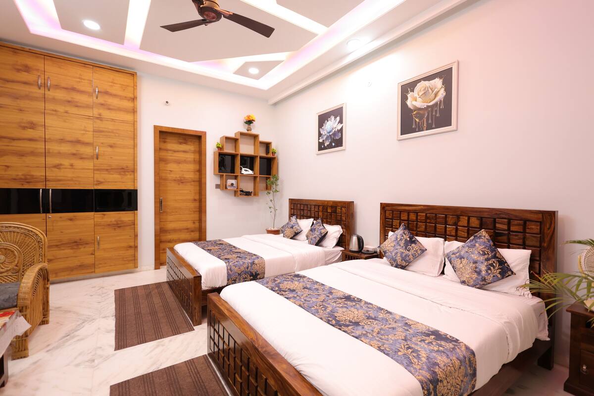 A hotel room with two beds, wooden headboards, a ceiling fan, a wooden wardrobe, a wicker chair, a small shelf with decorative items, and a framed picture on the wall.